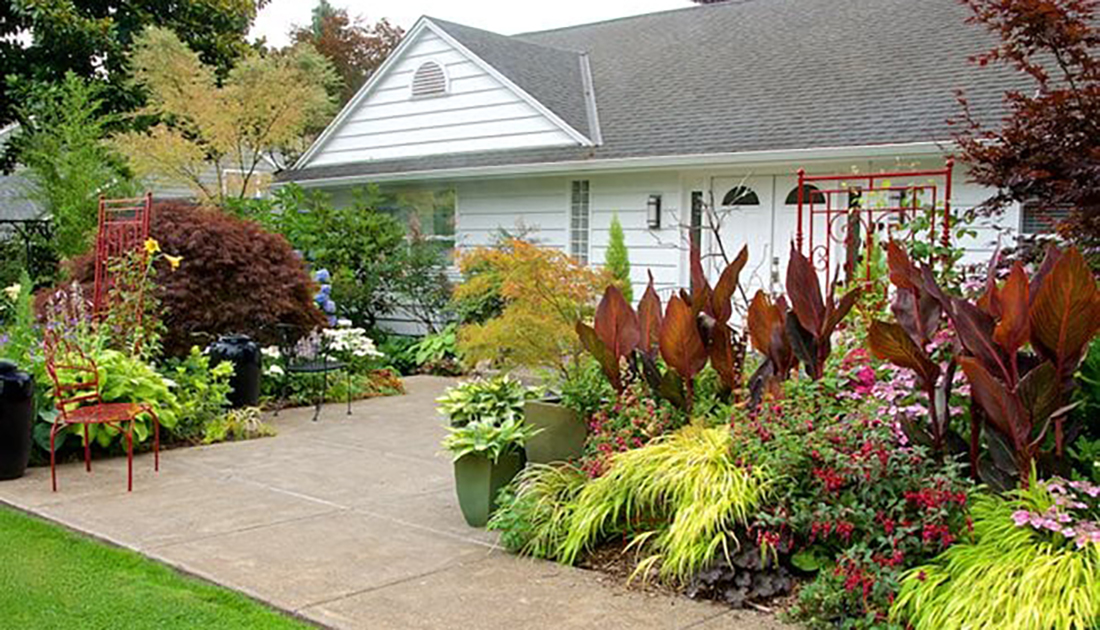 A lush residential garden in front of a white home, featuring a variety of colorful plants, flowers, and a red arbor, with a brick pathway leading to the entrance.